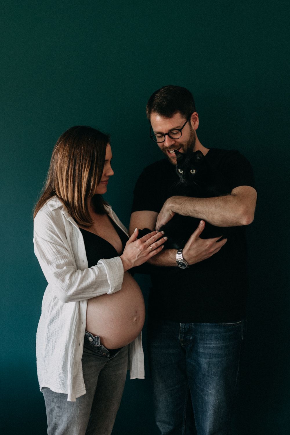Expectant parents with a black cat pose for a maternity portrait against emerald background.