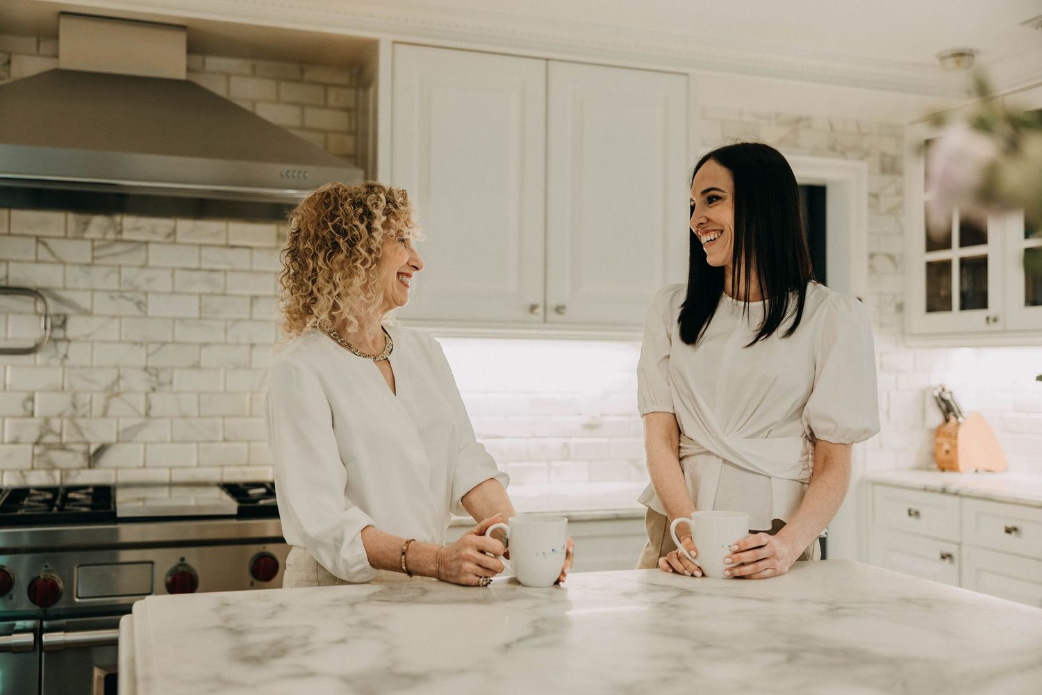 Two women stand at a marble kitchen counter having a conversation in a bright modern white kitchen.