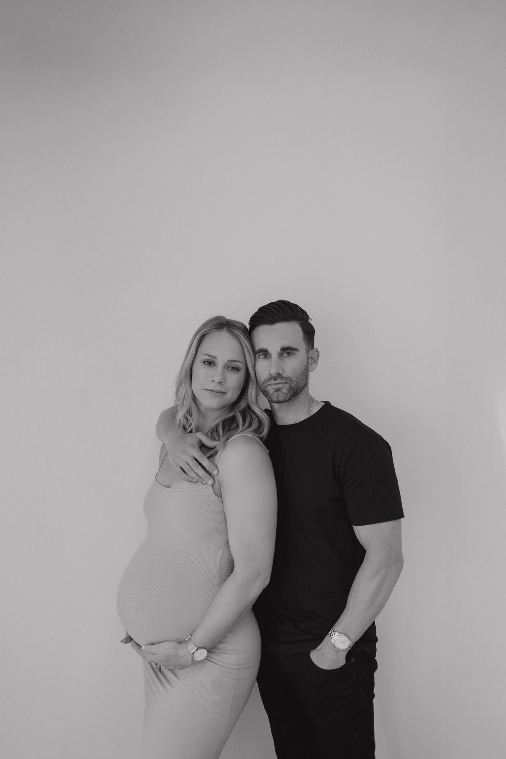 Black and white maternity portrait of couple embracing against plain wall.