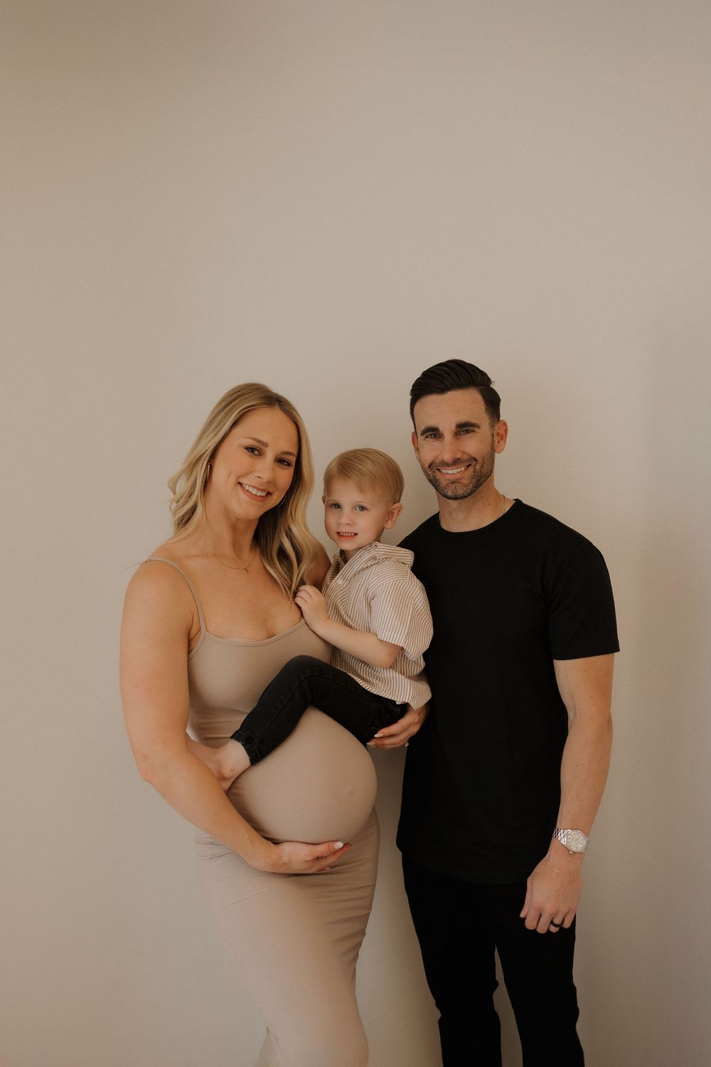 Family maternity portrait with expecting mother in beige dress against neutral backdrop.