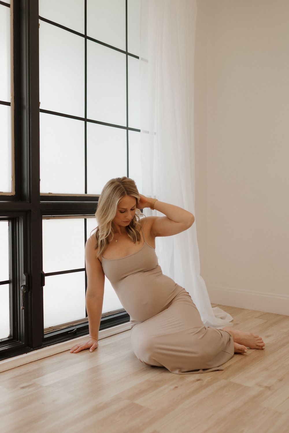 Expecting mother in beige dress poses elegantly by large window during maternity session.
