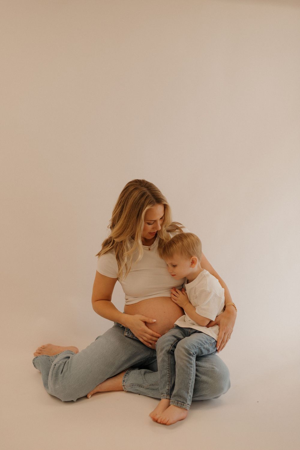 Intimate maternity photo of pregnant woman in casual attire bonding with toddler on neutral background.