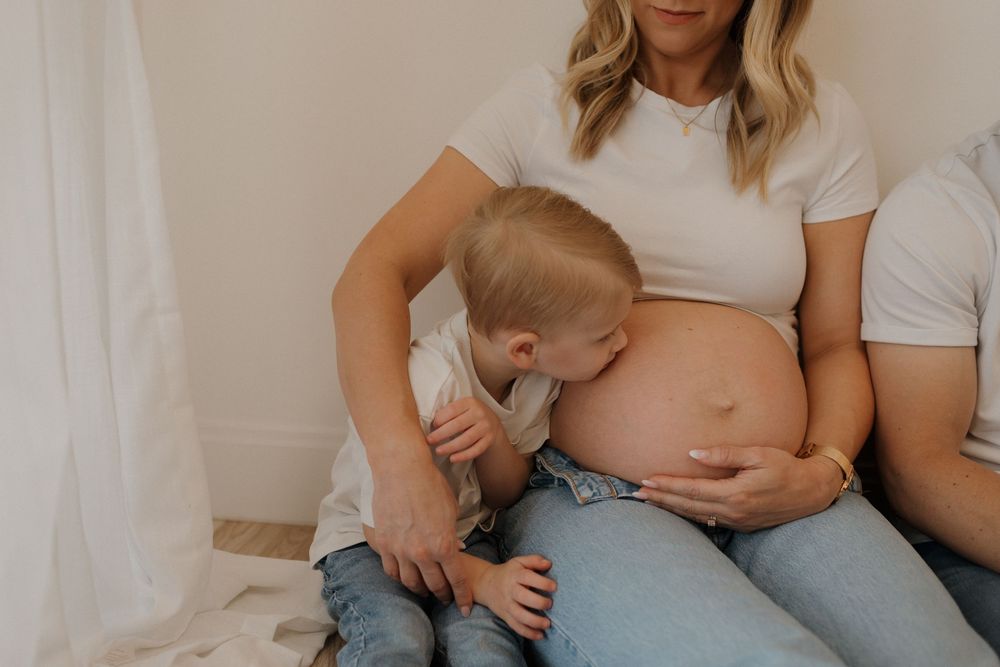 Candid maternity moment with toddler kissing pregnant belly while mother sits in casual attire.