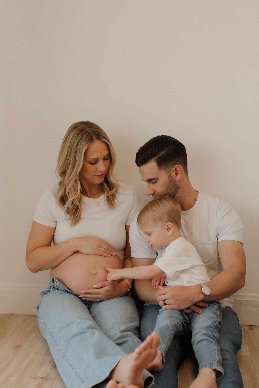 Candid family moment with toddler touching pregnant belly while parents sit on wooden floor.