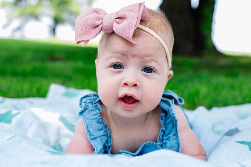 Baby in blue outfit with pink bow lying on white blanket outdoors.