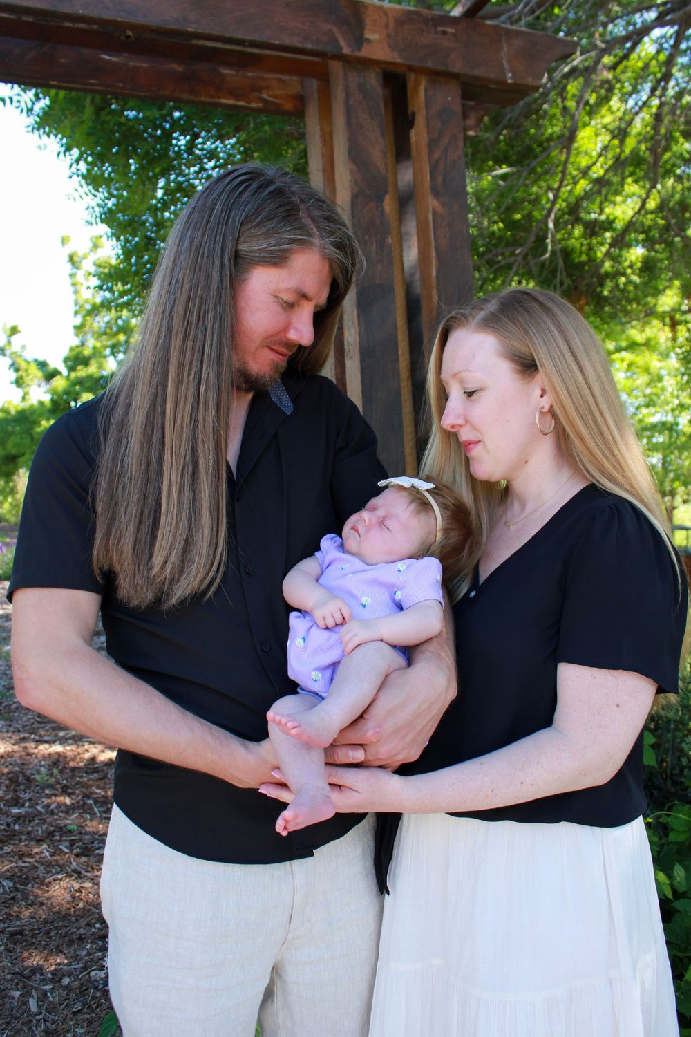 Couple in black shirts holding a sleeping baby in purple outfit under a wooden pergola outdoors.
