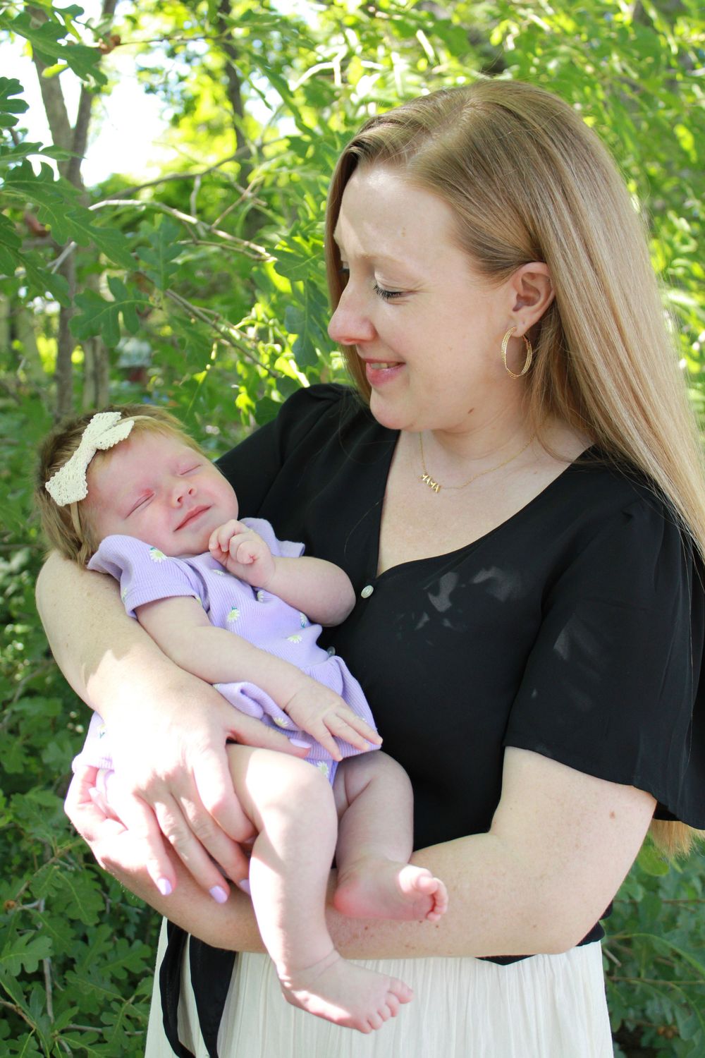 A woman in a black top smiles while holding a sleeping baby in a purple outfit outdoors among leafy green trees.