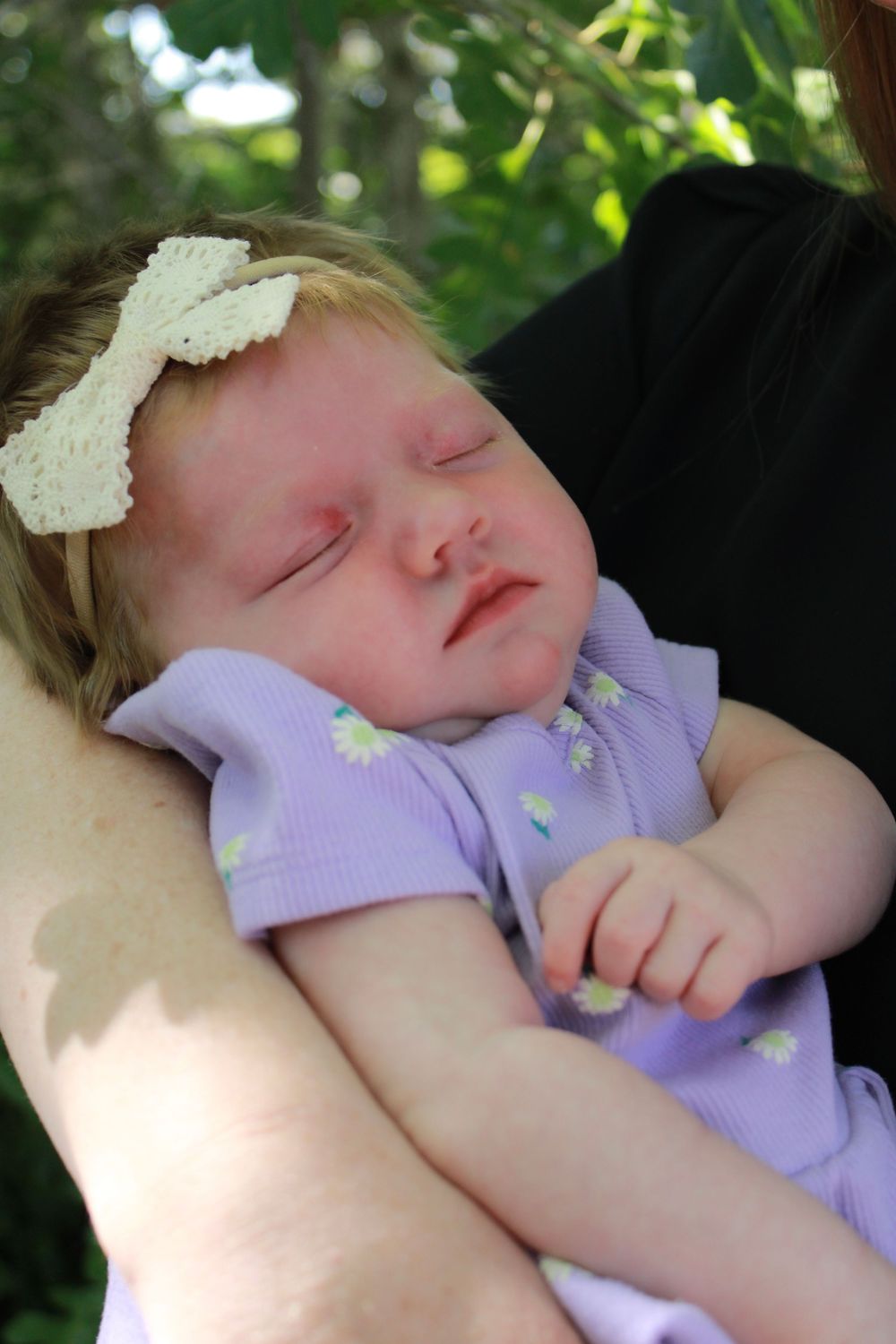 Sleeping baby in purple outfit wearing white bow headband resting peacefully against caregiver.