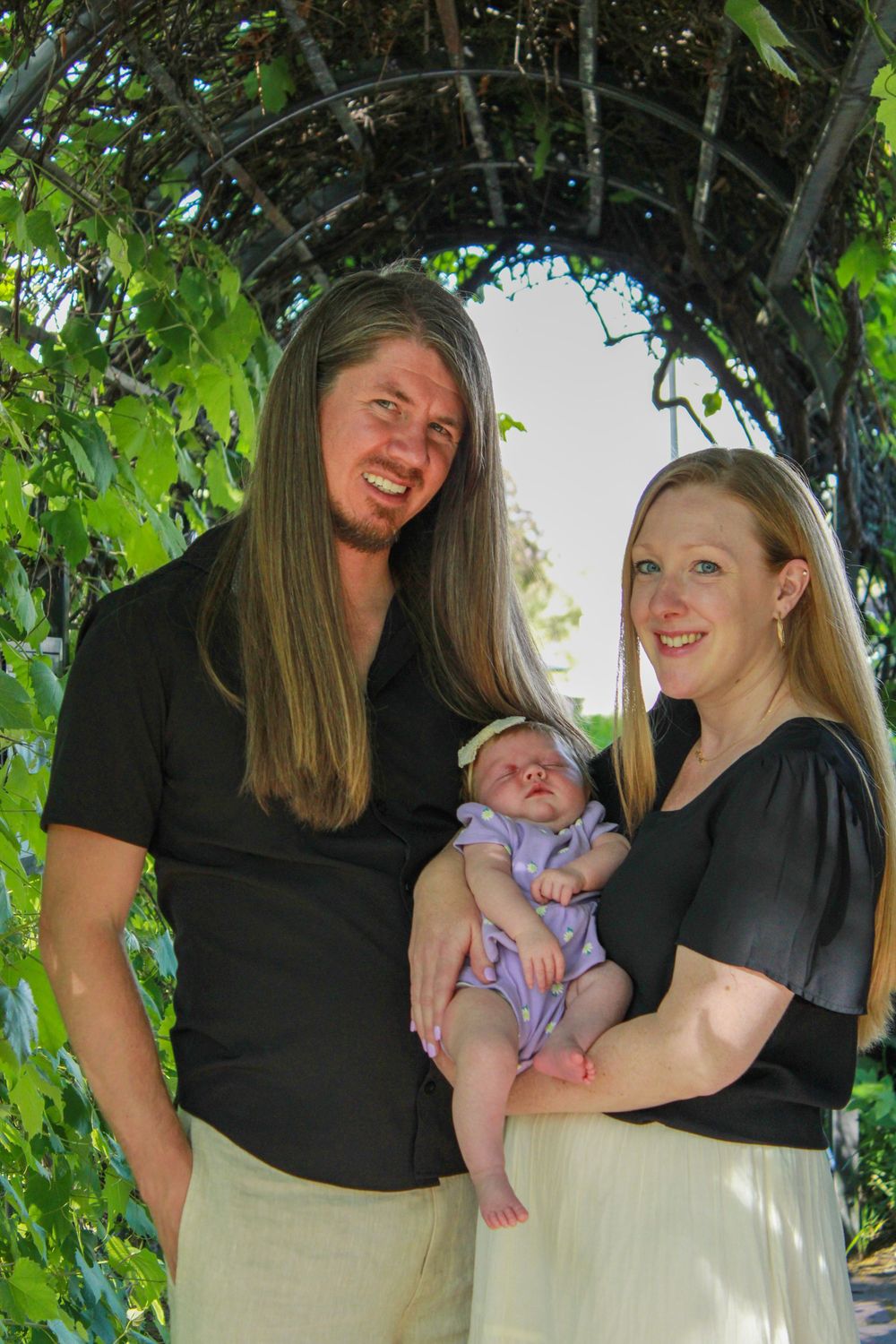 Family portrait under a vine-covered arch with newborn baby in purple outfit.