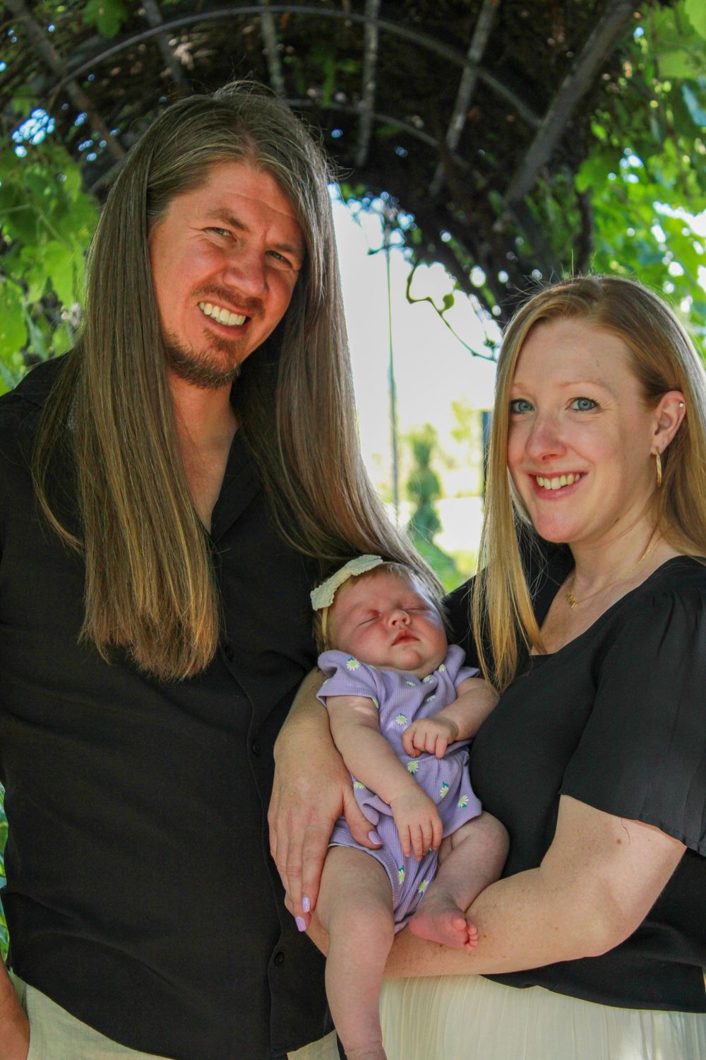 A family poses together outdoors with their newborn baby wearing purple under a green garden arbor.
