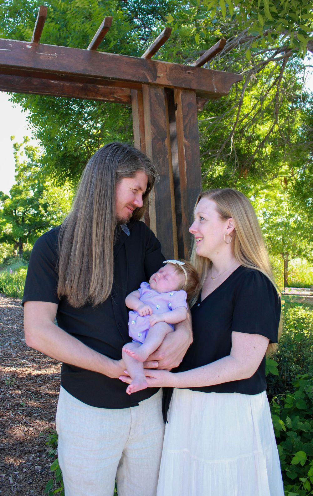 Two adults in matching black tops and white bottoms share a tender moment with an infant under a wooden pergola in a garden.