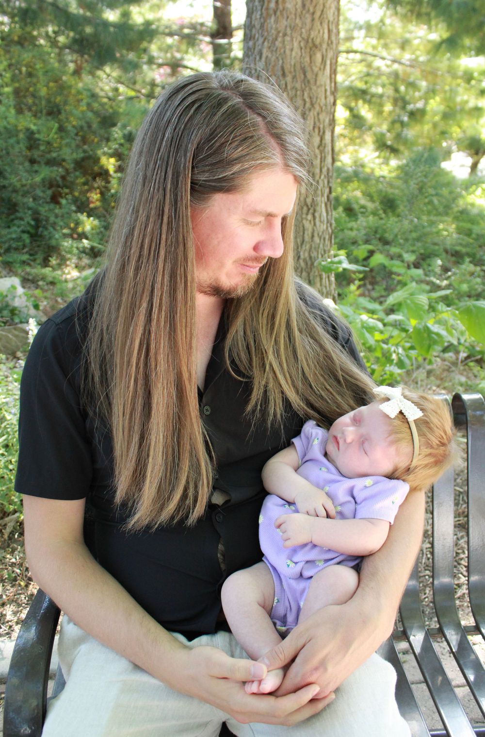 A parent tenderly holds a sleeping newborn wearing purple while sitting on a park bench surrounded by greenery.