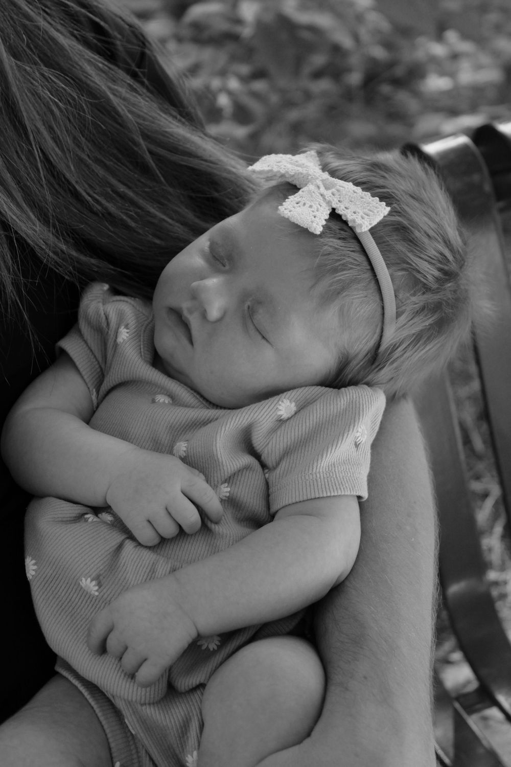 Black and white portrait of sleeping baby wearing white bow headband resting on someone's shoulder.