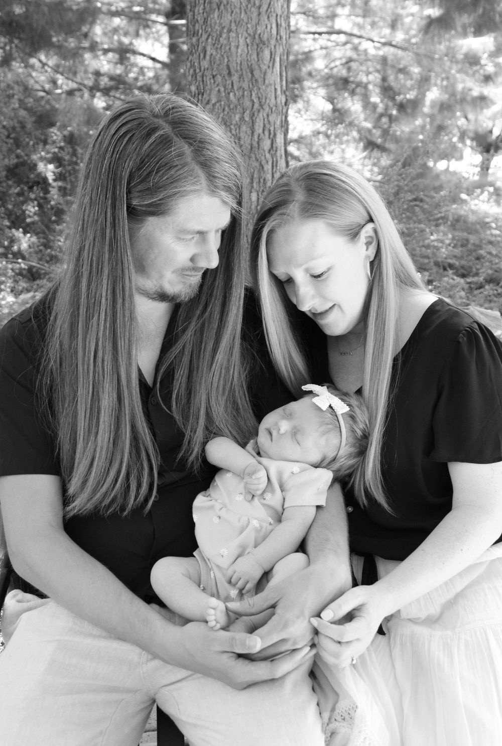 A black and white portrait of a couple cradling their sleeping newborn baby outdoors by a tree trunk.