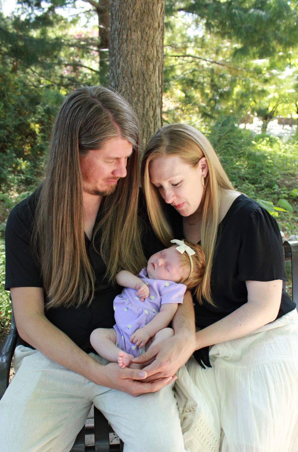 A couple sits outdoors holding their newborn baby dressed in purple while gazing lovingly at their child.