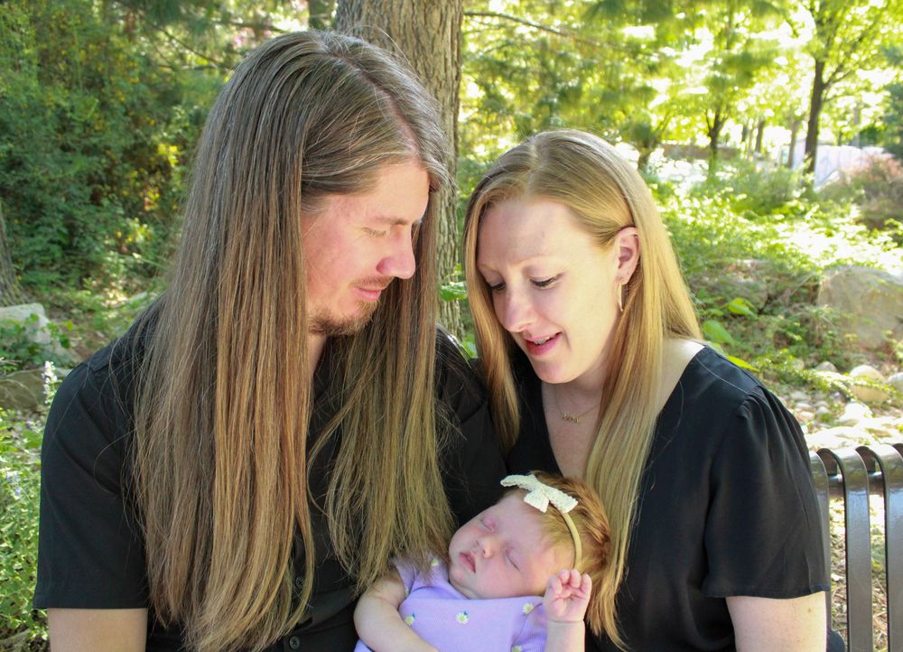 Two people in black tops looking down tenderly at a small baby wearing a gold headband outdoors in a garden setting.