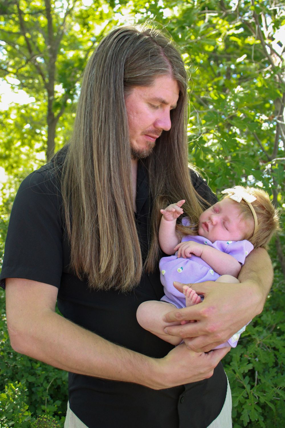 Parent in black shirt with long hair tenderly holding a sleeping baby wearing purple outdoors.