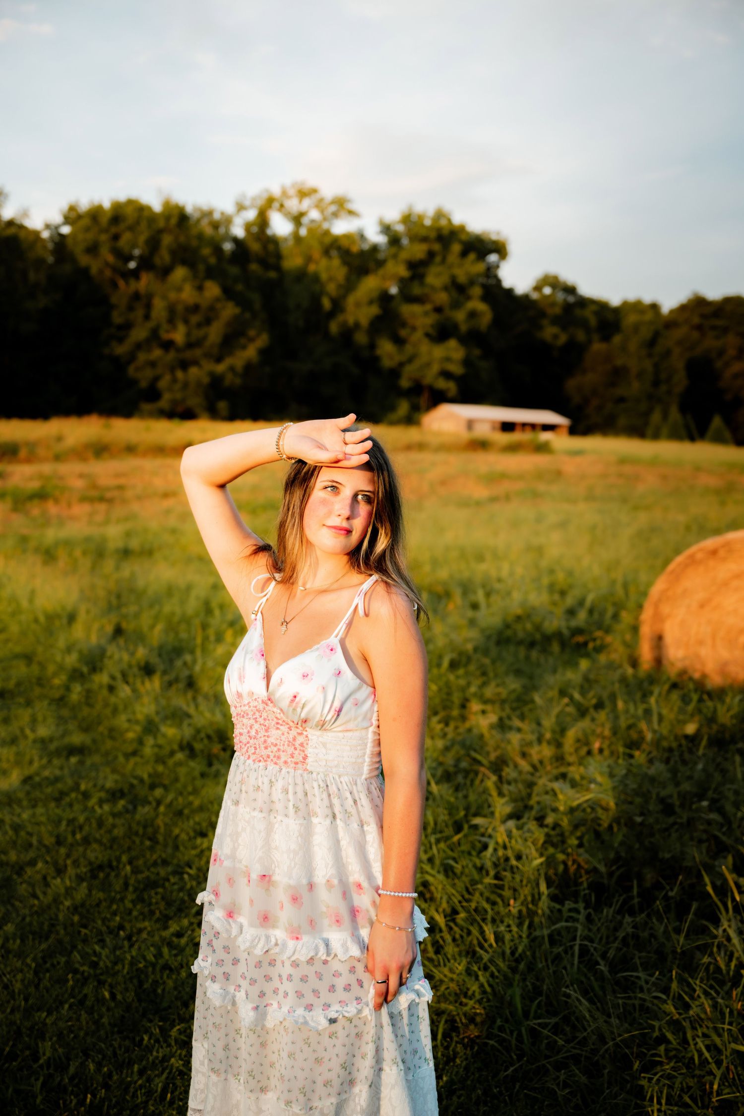 Young woman in white floral maxi dress poses in golden hour sunlight in a rural field with hay bales and trees.