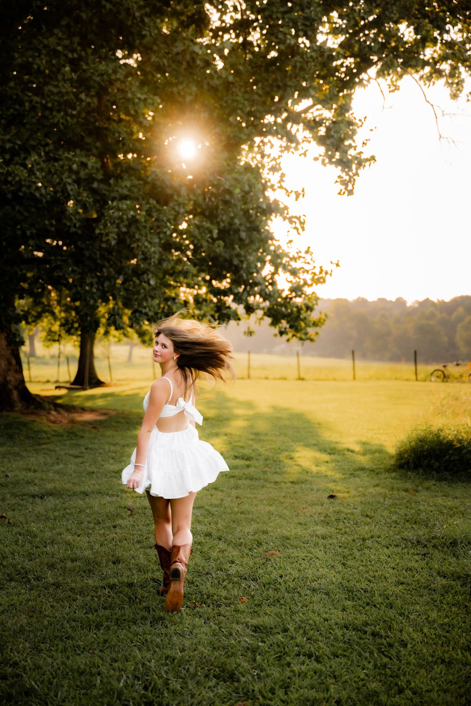 Young woman in white dress and cowboy boots twirling in a sunlit green field at golden hour.