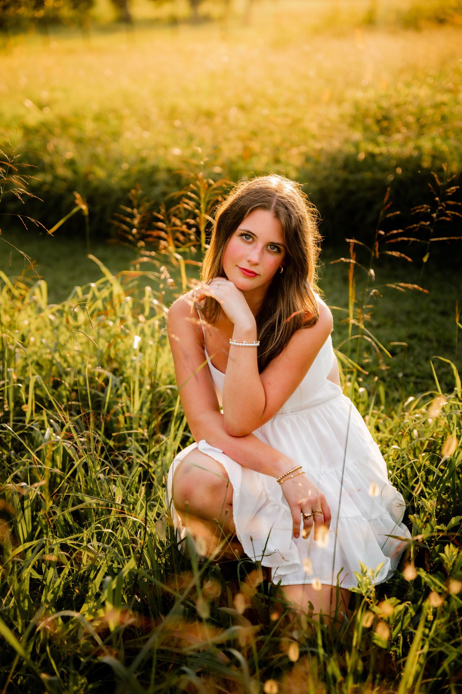 Young woman in white dress crouching in golden sunlit meadow with tall grass, wearing jewelry, golden hour portrait.
