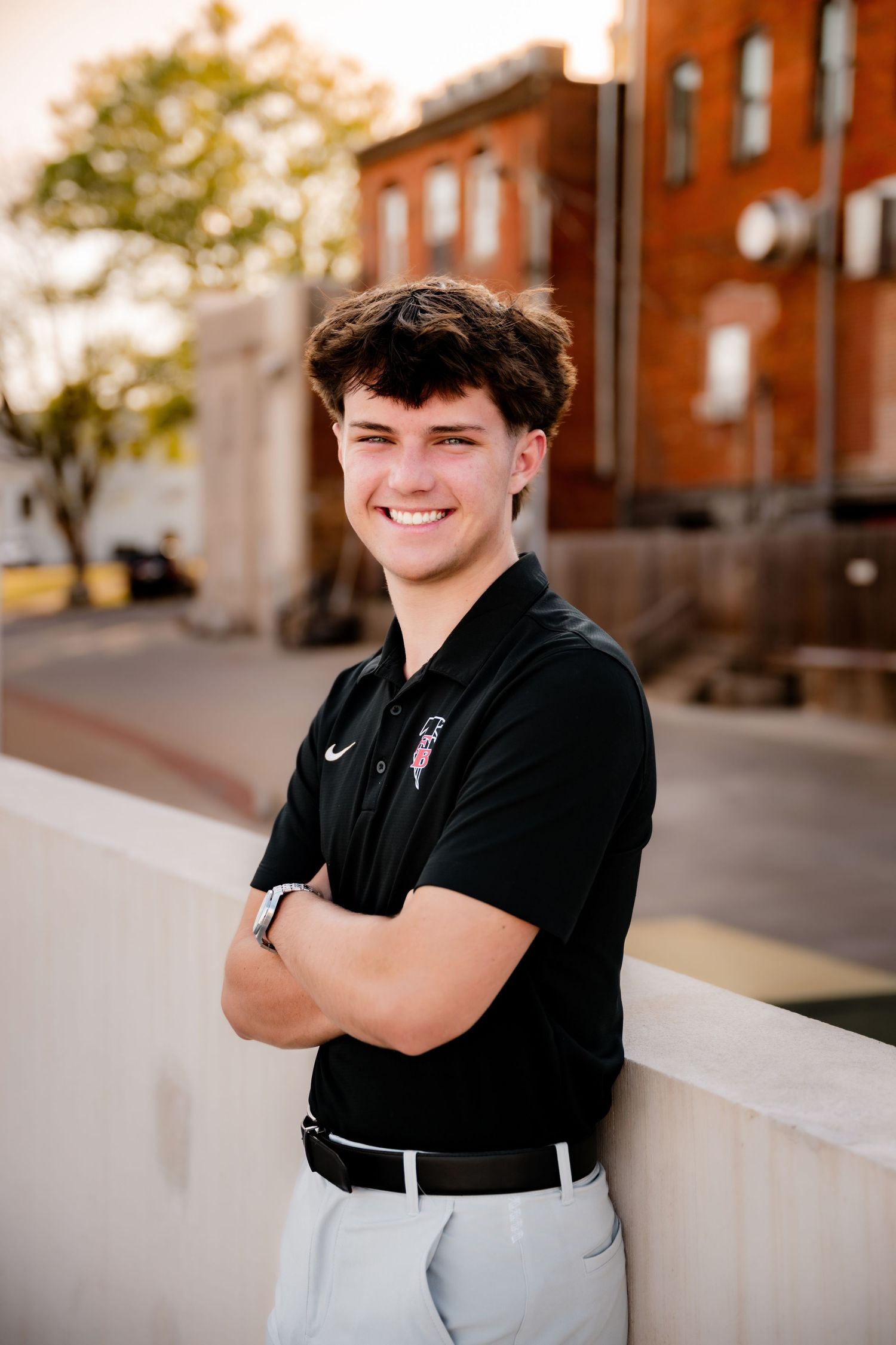 Smiling young man in black Nike polo with arms crossed, posing outdoors near brick buildings at golden hour.