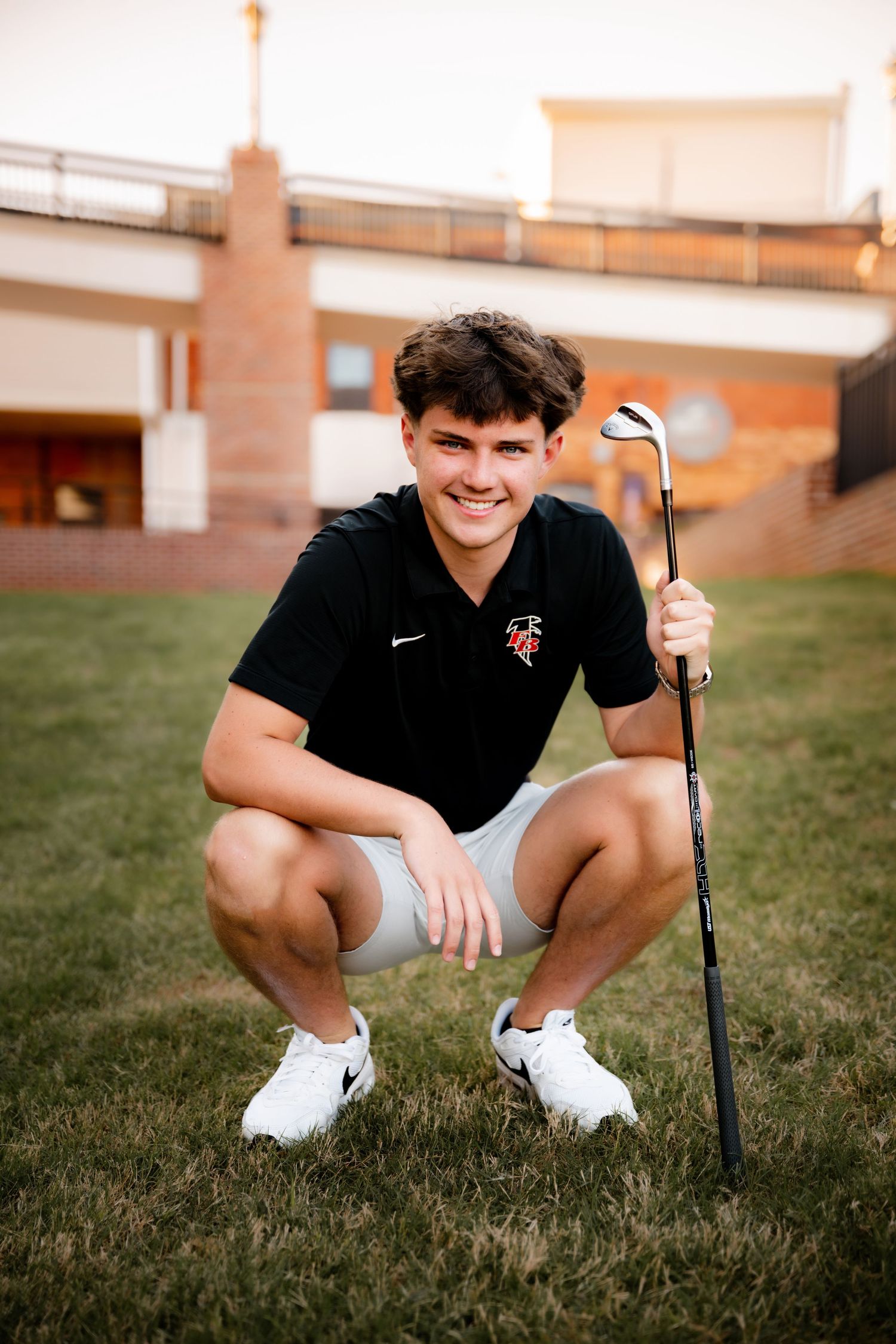Young male golfer crouching on grass, holding a golf club, wearing a black Nike polo and white shorts.