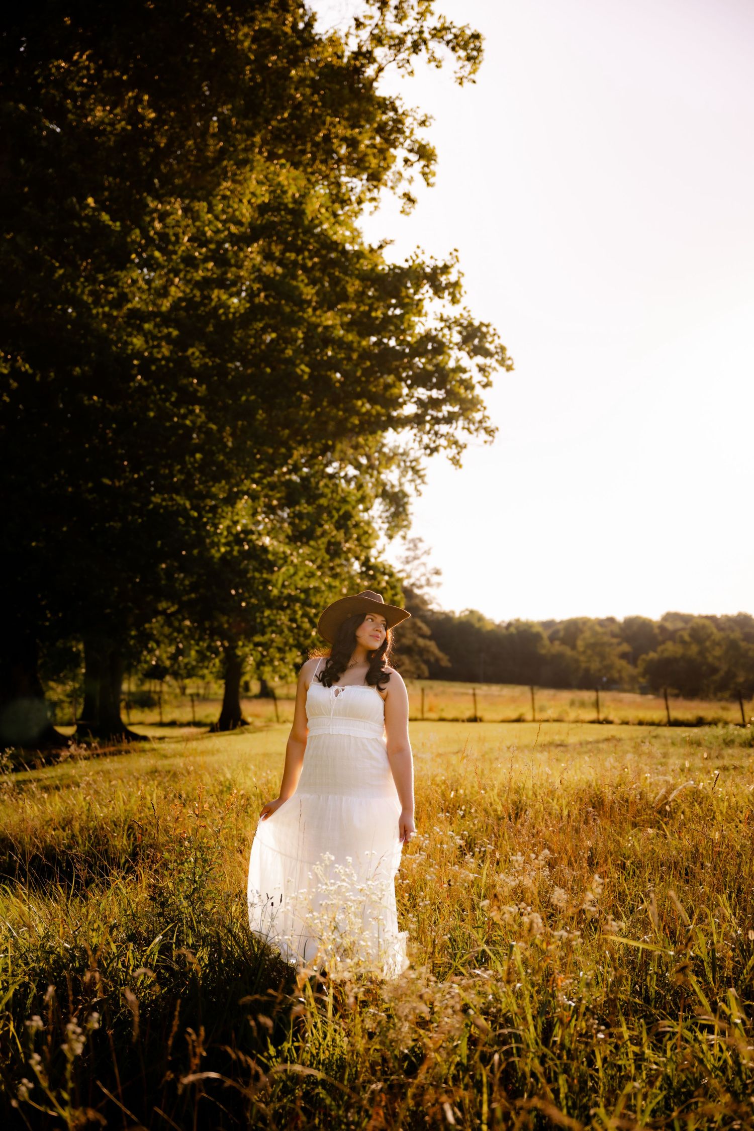 Woman in white lace dress and hat standing in sunlit meadow with large trees in golden hour light.