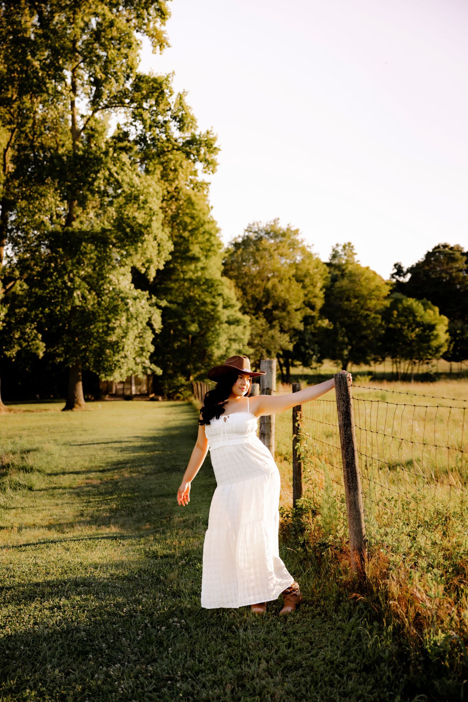 Pregnant woman in white dress and cowboy hat leaning on rustic fence in golden hour sunlight.
