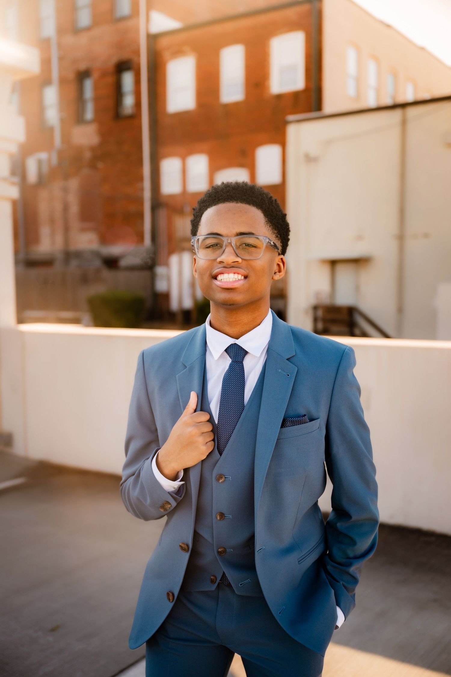 Professional in navy blue suit posing against brick building at sunset.