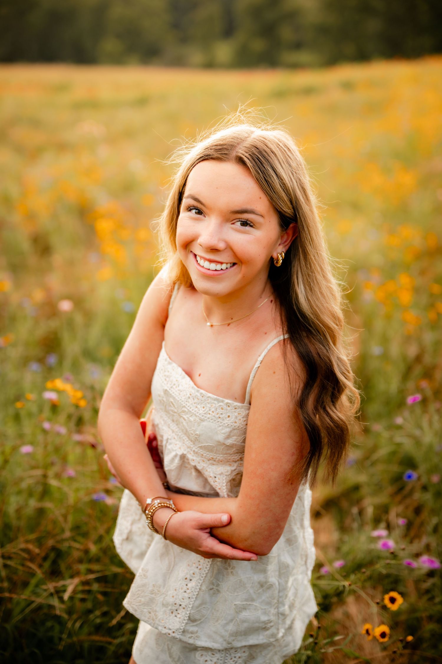 Smiling teenage girl in white dress standing in colorful wildflower field with yellow and purple blooms.