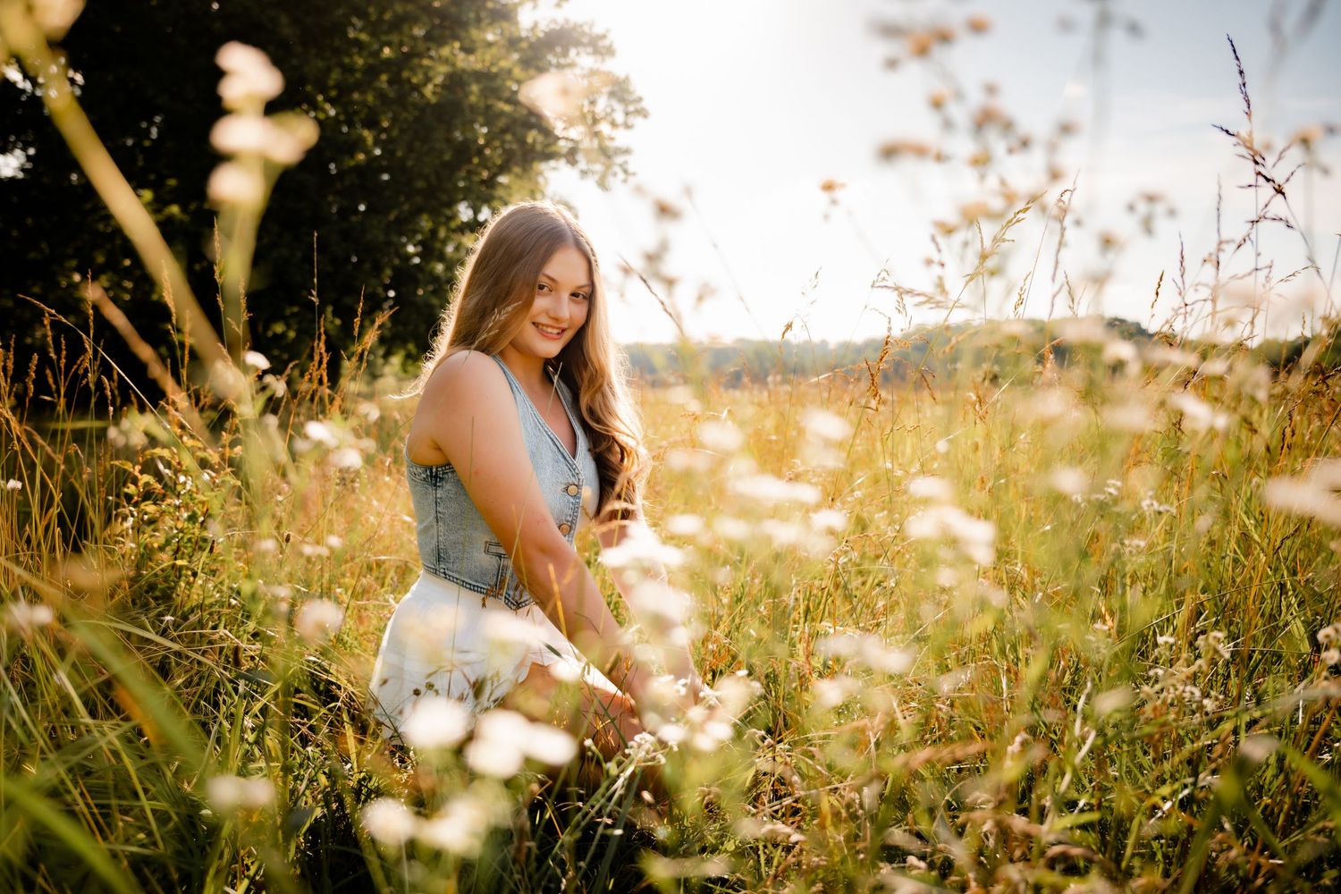 Young woman smiling in a sunny wildflower meadow, holding flowers, wearing a blue top and white skirt.