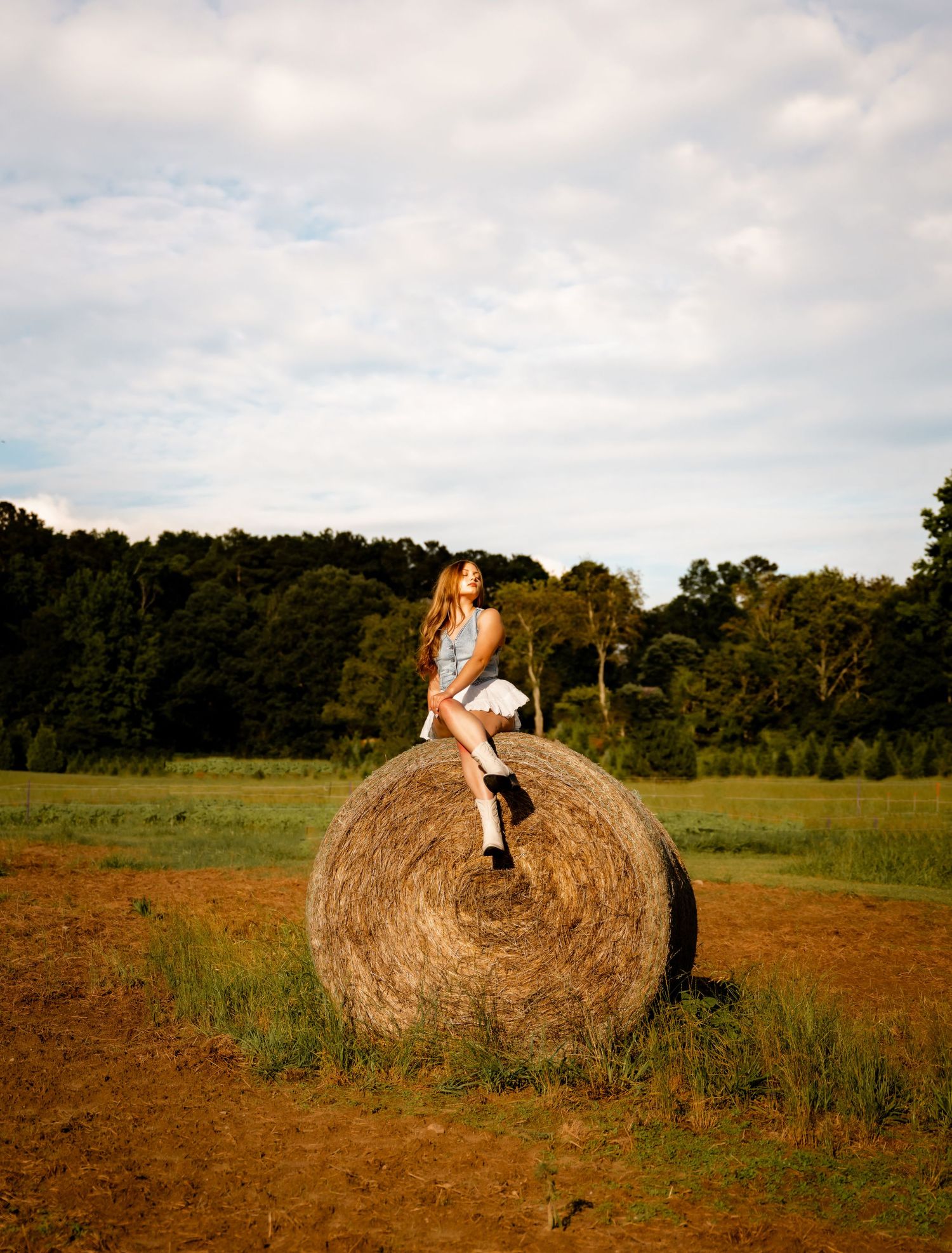 A young woman in boots sits atop a large round hay bale in a golden farm field with trees in the background.