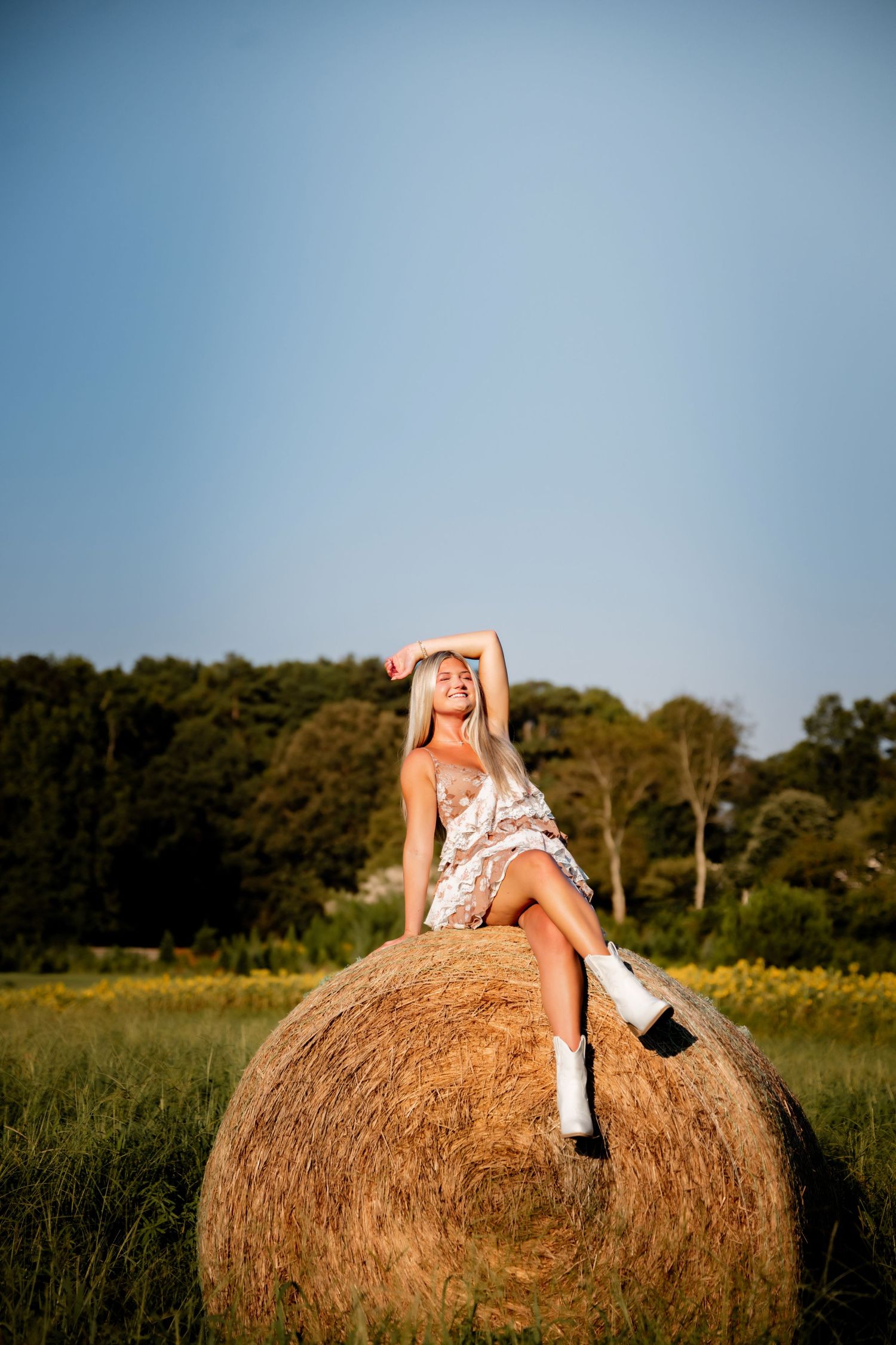 Blonde woman in floral dress and white boots sitting atop a round hay bale in a green field under blue sky.