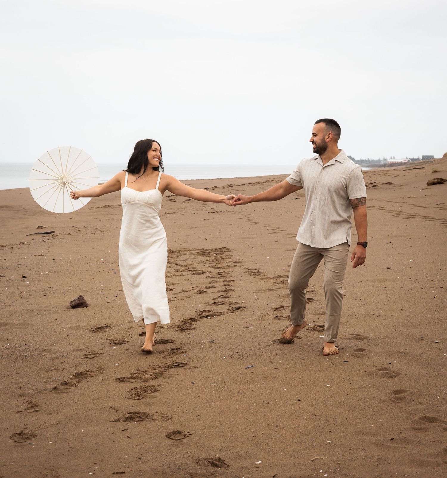 A couple in light-colored clothing dances playfully together on sandy dunes at sunset.