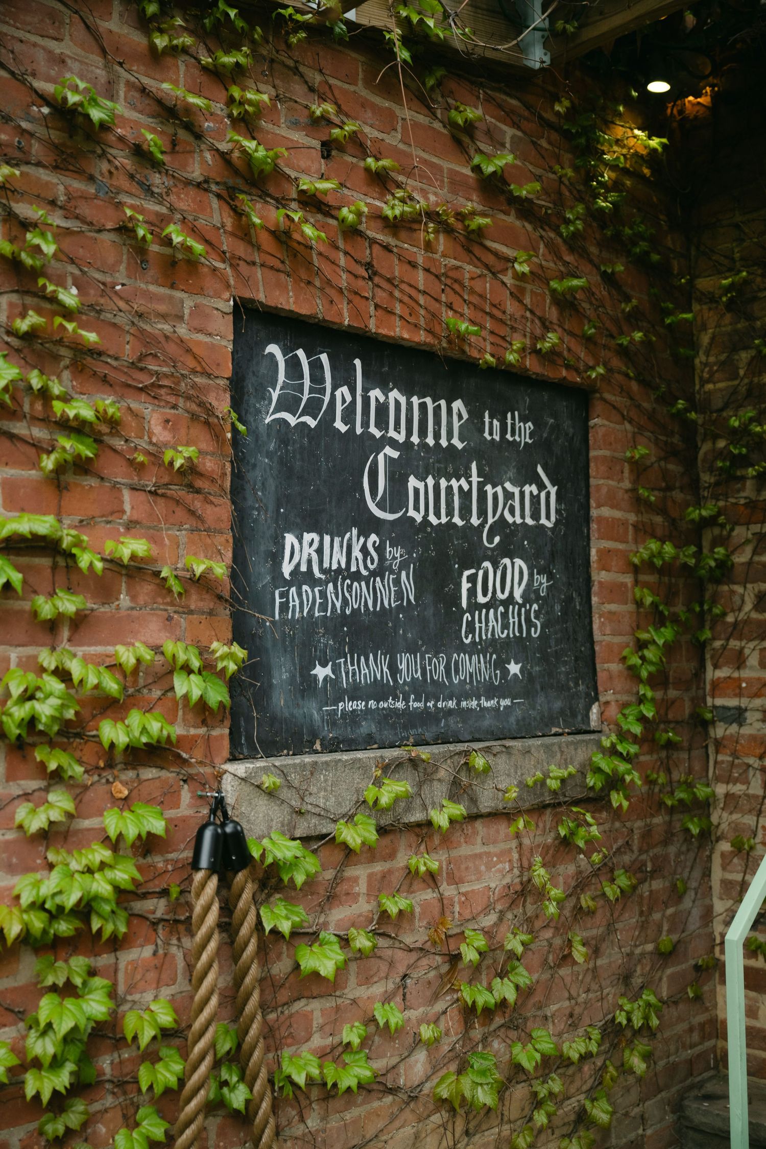 A rustic courtyard welcome sign on ivy-covered brick wall advertising drinks and food pavilion.