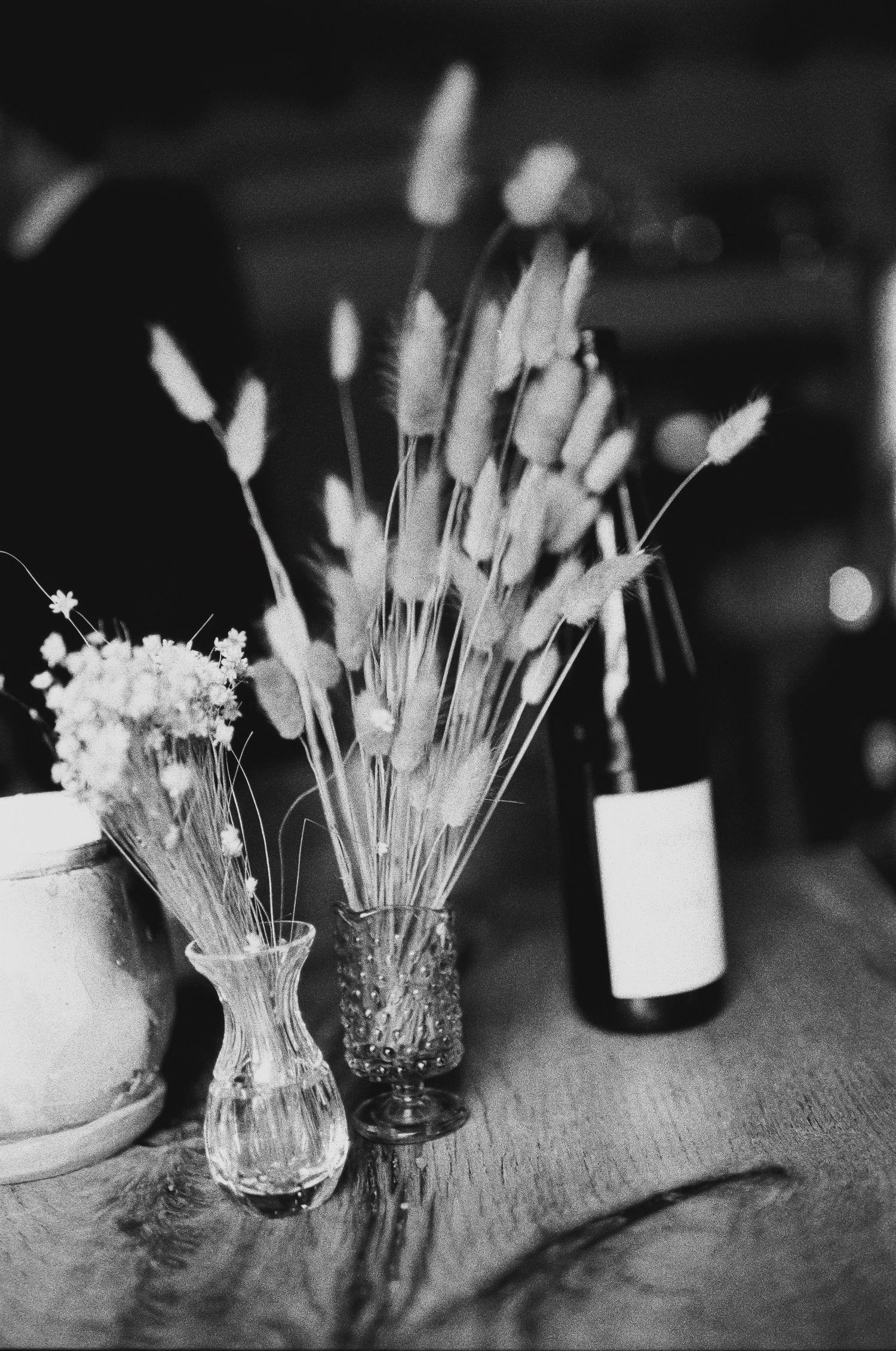 Black and white photo of dried bunny tail grass arrangements in glass vases with a wine bottle on wooden table at Fadensonnen in Baltimore