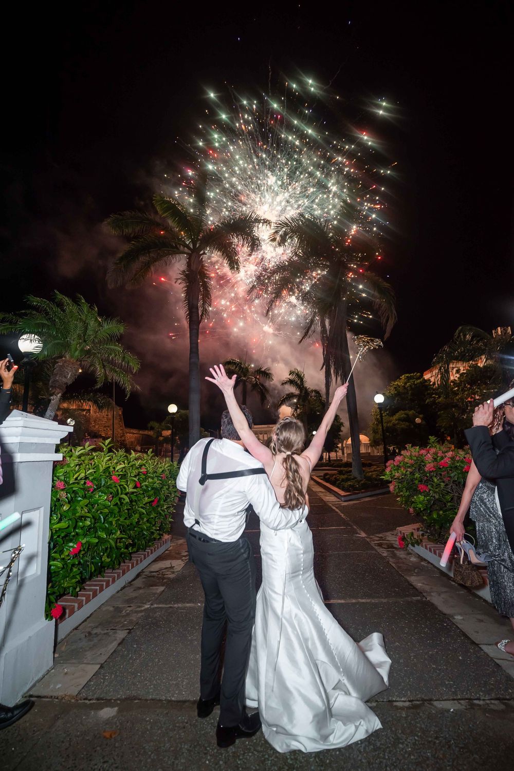A couple celebrates under spectacular fireworks display at night with palm trees silhouetted in a tropical wedding setting.