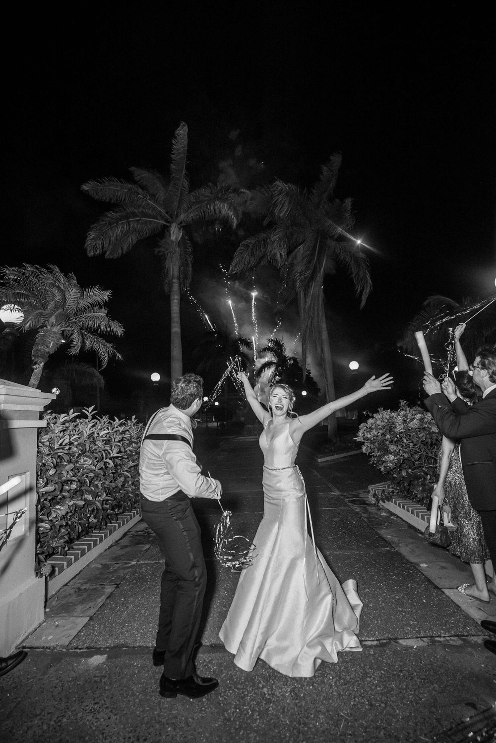 Black and white nighttime celebration photo with palm trees and sparklers creating a festive atmosphere outdoors.