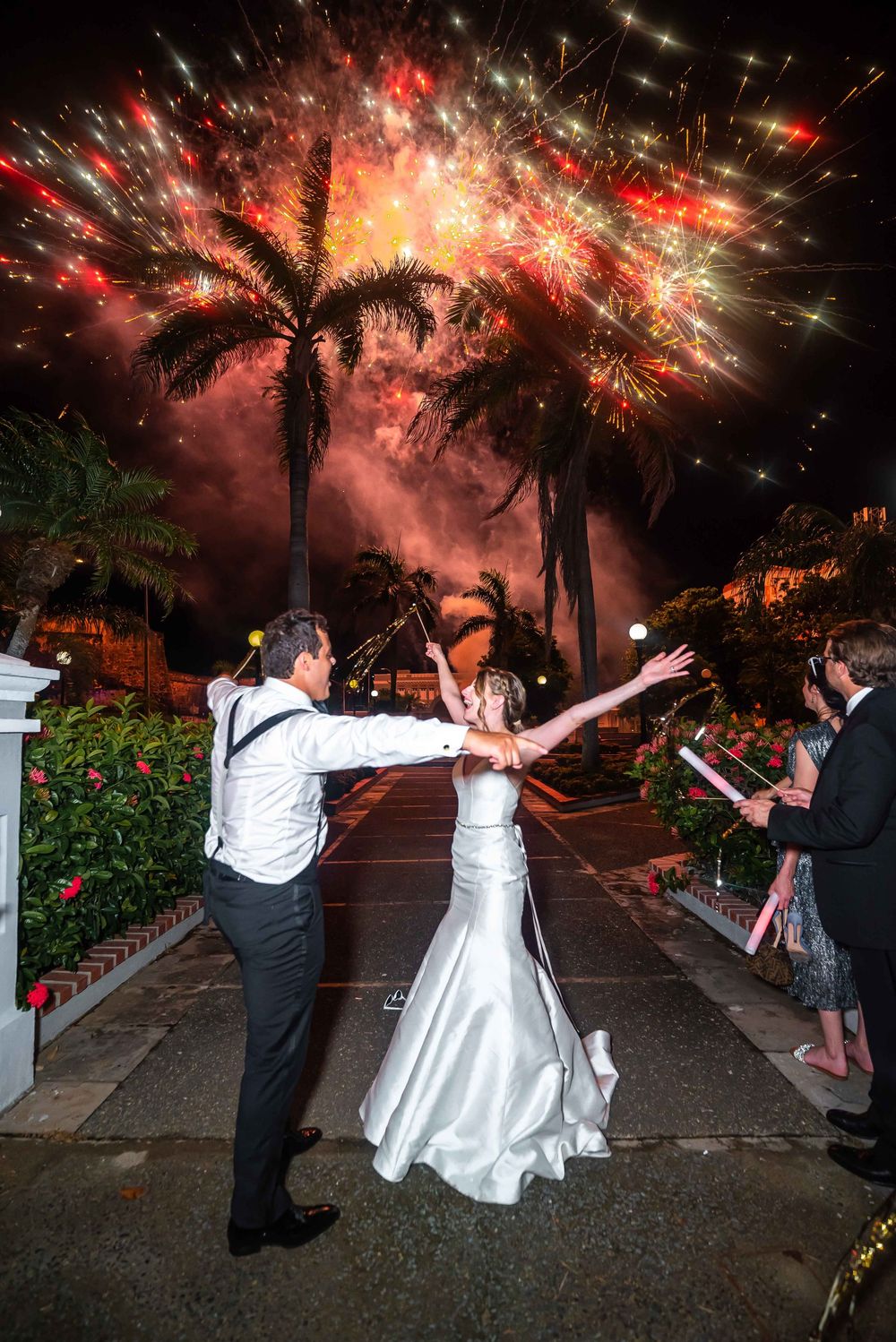 Wedding celebration at night with couple dancing under palm trees and colorful fireworks display in the tropical sky.