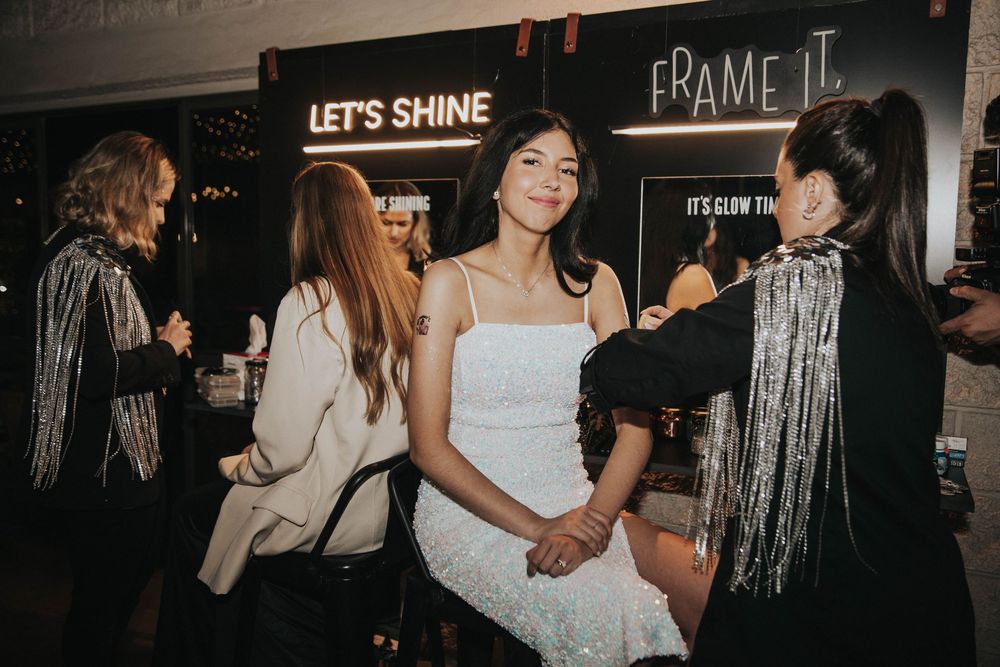 Invitados con ropa de lentejuelas y vestidos, socializan en fiesta nocturna con señalización iluminada, ideal para stand de glitter