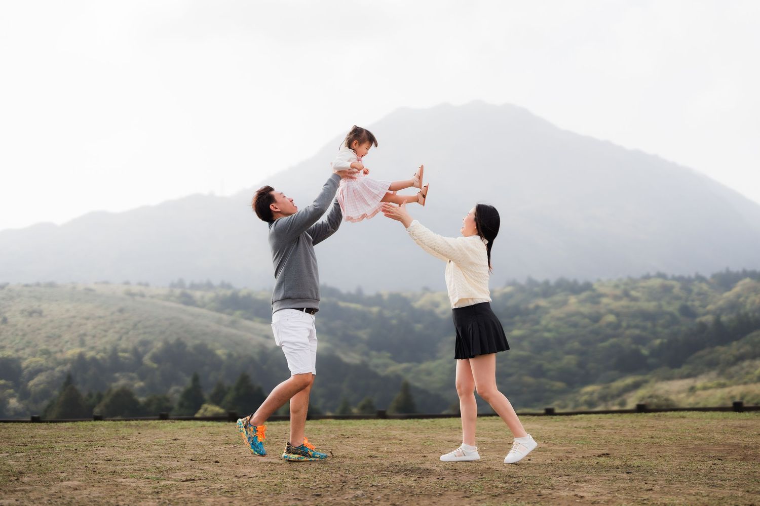 Parents toss their child in the air while playing together on a grassy hilltop with mountains in the background.