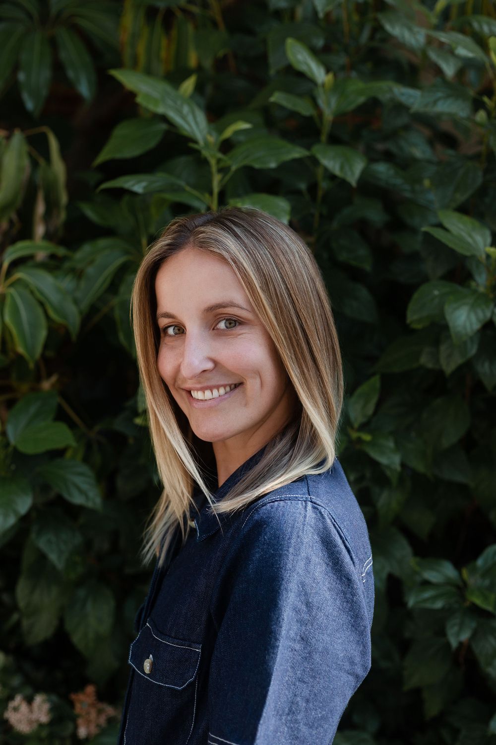 Casual outdoor headshot in denim jacket with green leaves — natural light portrait by Adrian Rudd Kahn.