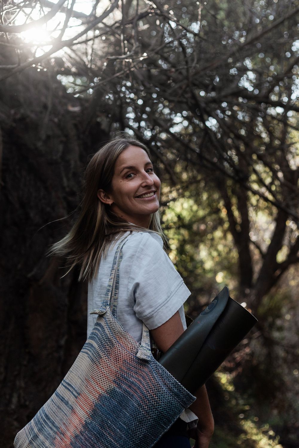 Woman walking through forest with yoga mat in hand — natural light wellness photography by Adrian Rudd Kahn.