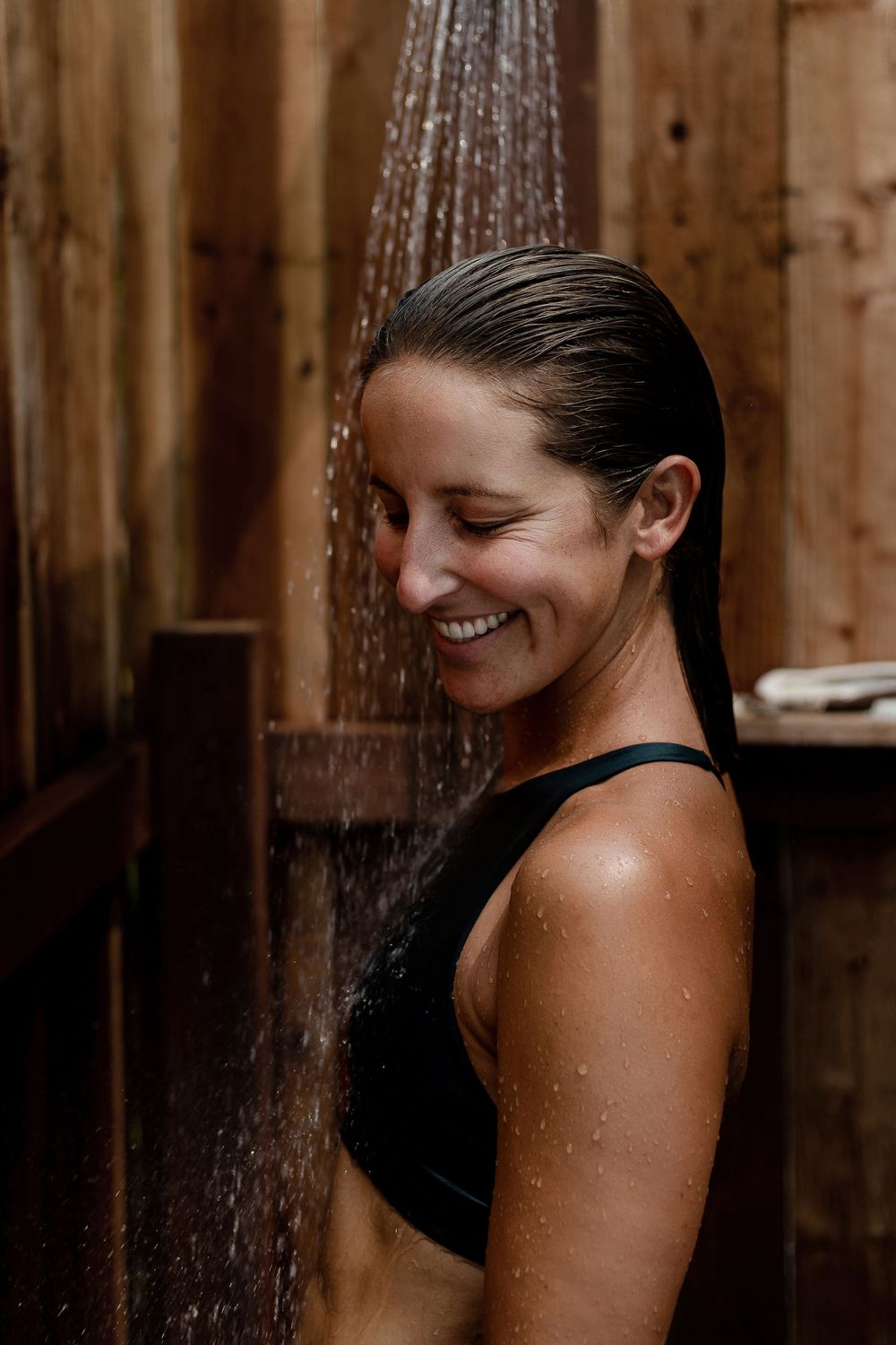 Smiling woman enjoying open-air shower in wood enclosure — wellness lifestyle photo by Adrian Rudd Kahn