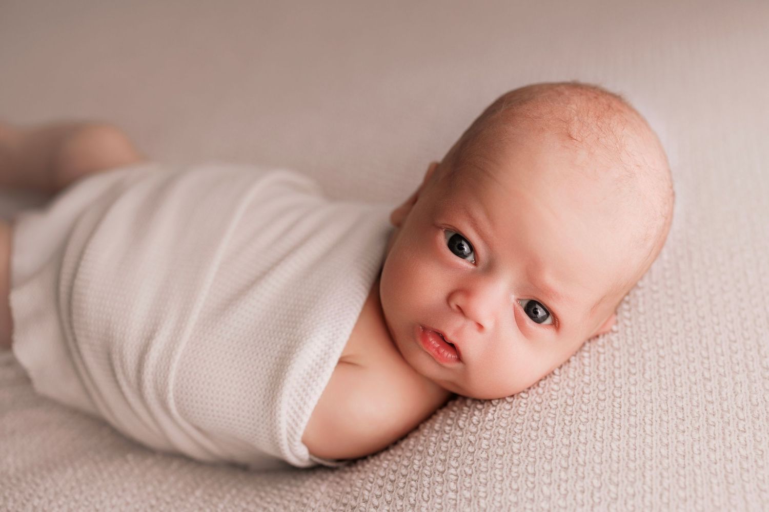 Young child in striped shirt gently touching a newborn baby wrapped in white clothes on bed.