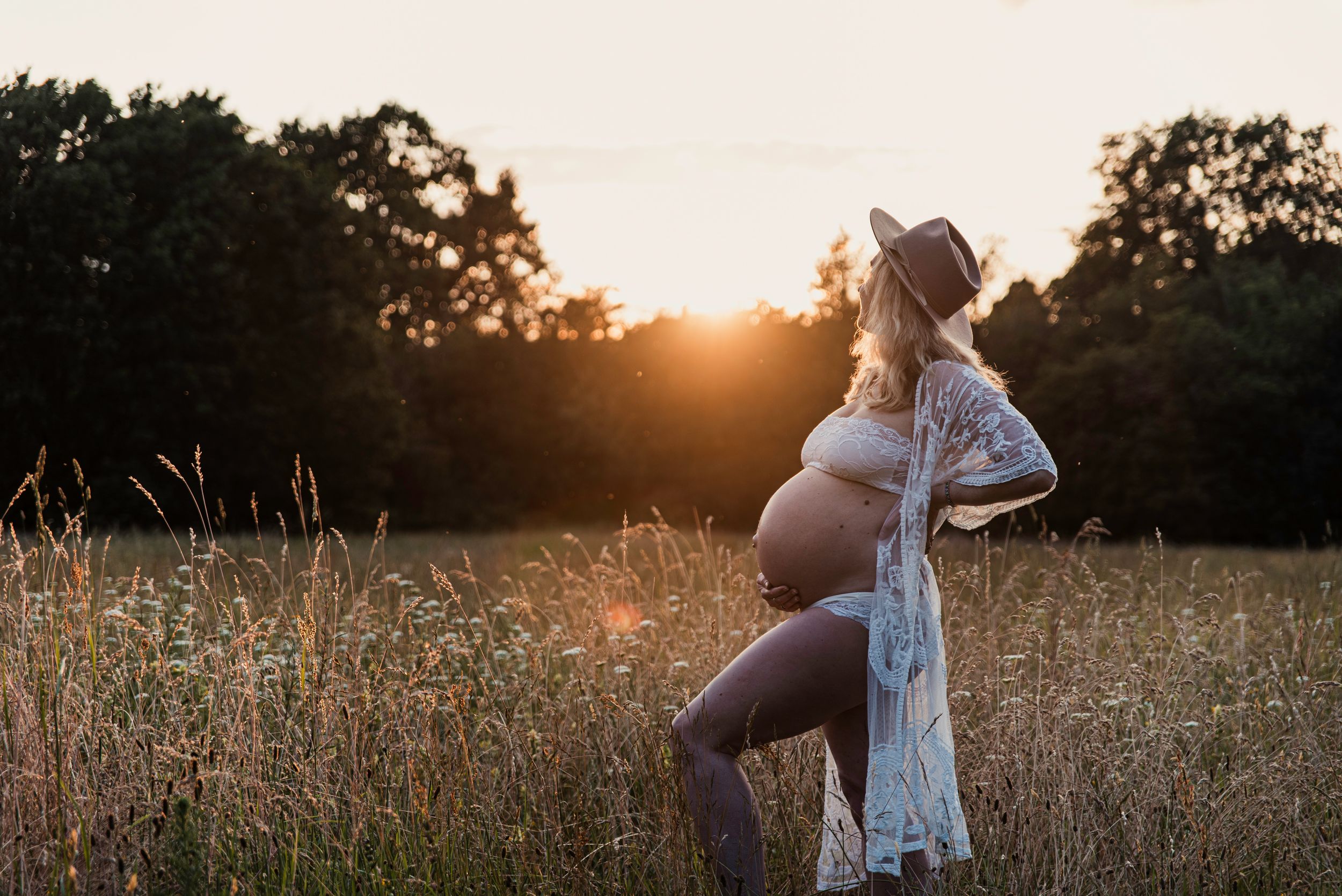 Silhouette wearing a lace dress and hat stands in a wildflower meadow during golden sunset.
