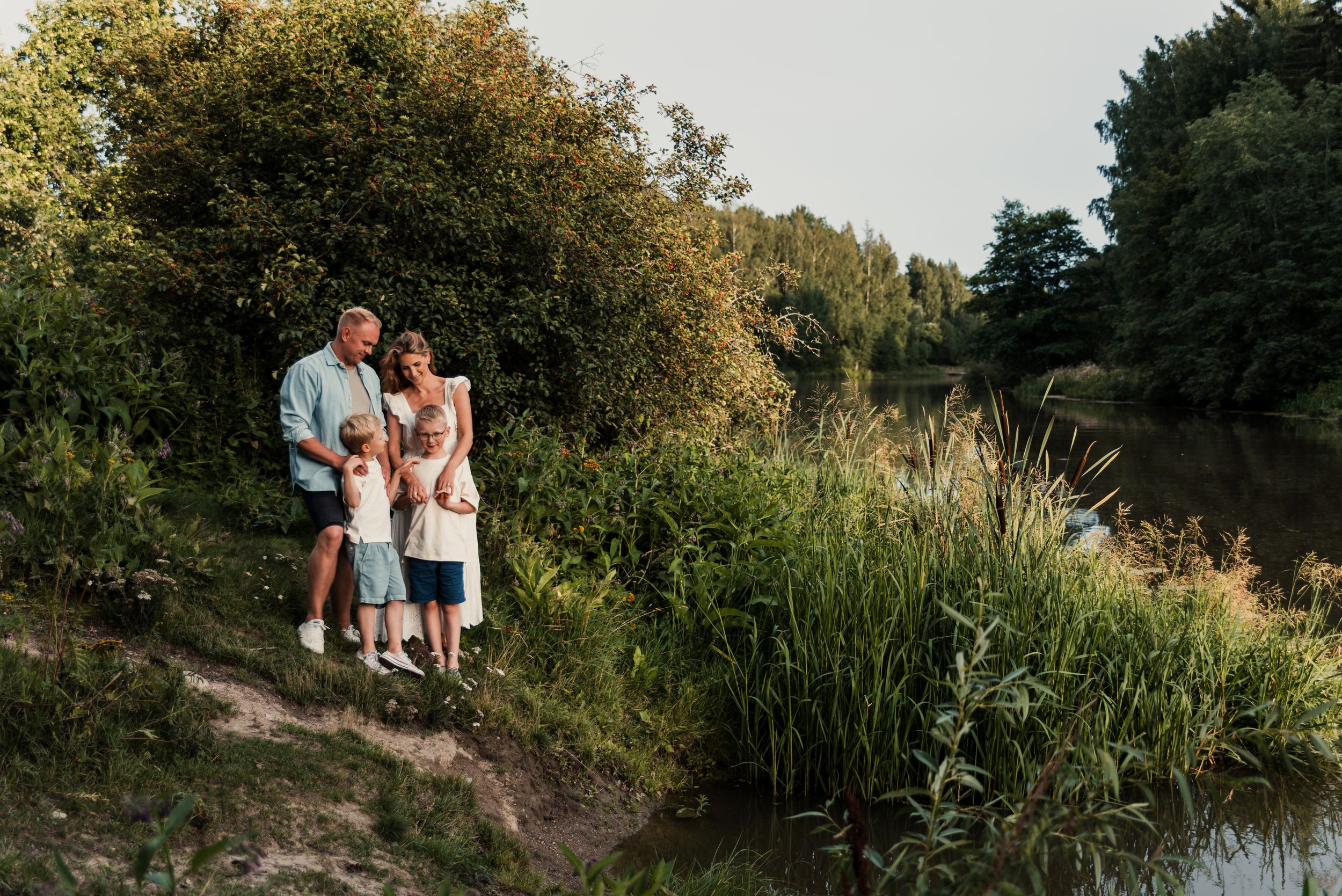A small group stands together on a grassy riverbank during a summer evening surrounded by tall reeds and trees.