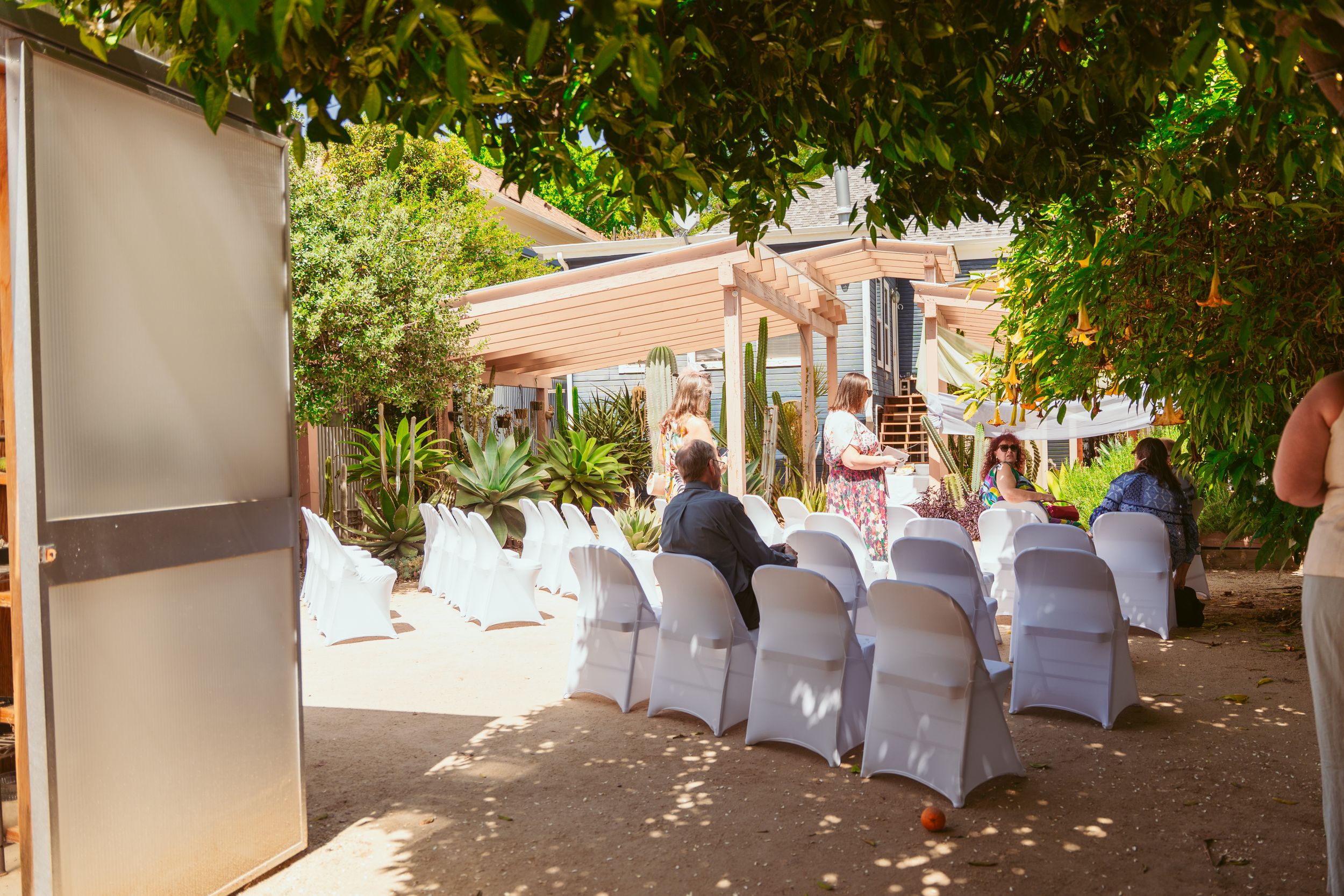 Outdoor wedding reception venue with white chair covers and dappled sunlight filtering through overhead greenery.