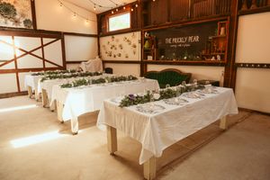 Rustic indoor venue setup with long tables covered in white linens and natural greenery decor.