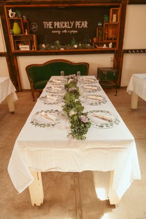 A formal dining table set with white linens and greenery garland under a sign reading 'The Prickly Pear'.
